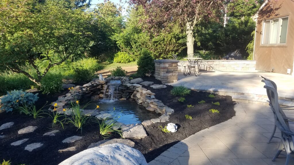 Image of an outdoor living space with a small artificial lake surrounded by stone, plants, trees, and stone hardscaping